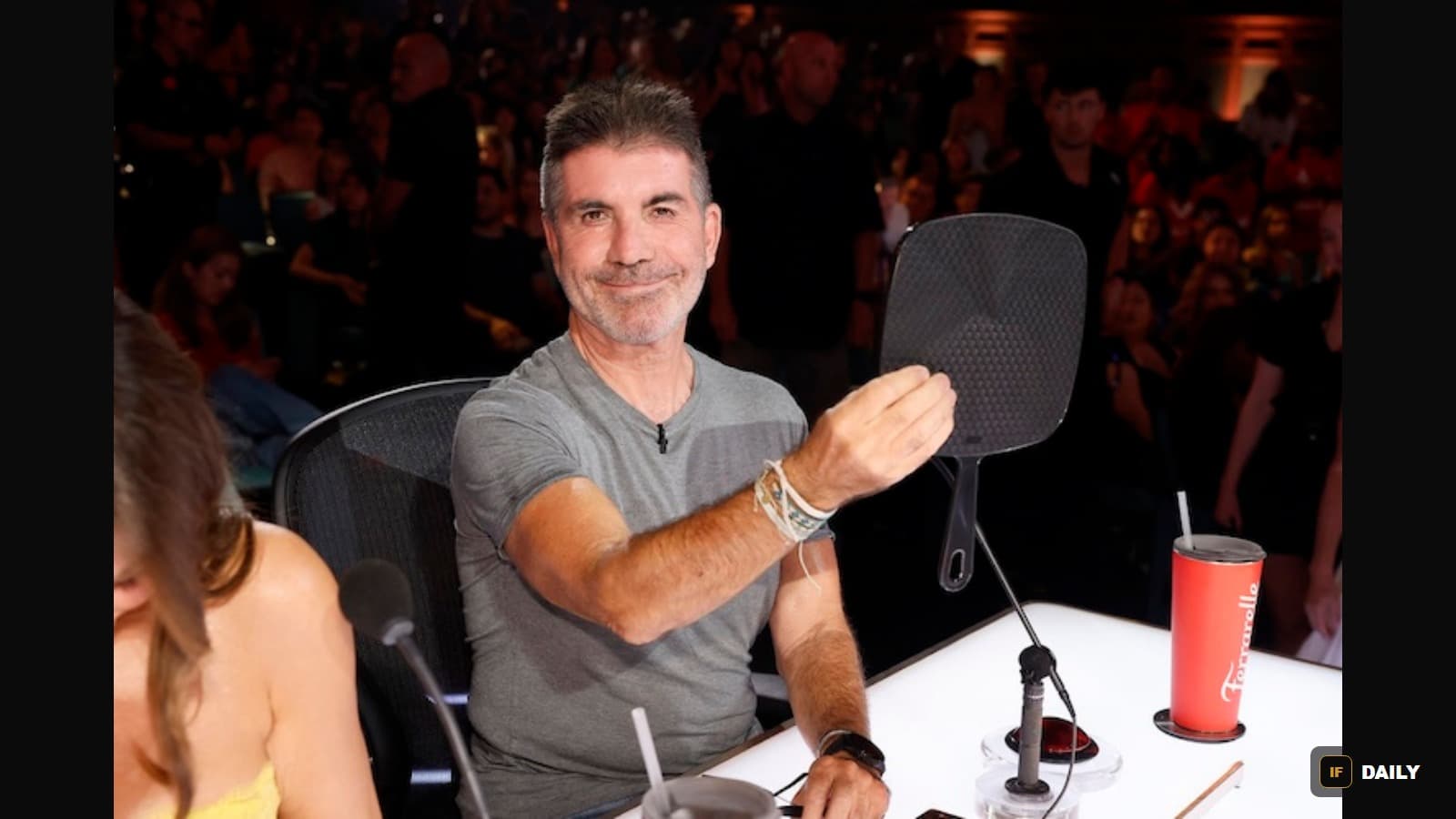 Simon Cowell seated at the AGT judges'' table holding a paddle in front of a live studio audience