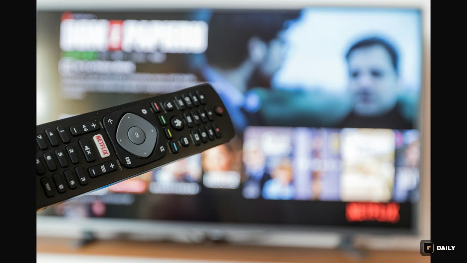 A TV remote control with a red Netflix button, held in front of a blurred television showing a streaming interface
